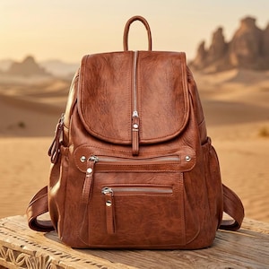 May include: A brown leather backpack with multiple zipped compartments and a top handle. The bag has a flap closure and adjustable shoulder straps. The backpack is on a wooden surface with a desert landscape in the background.