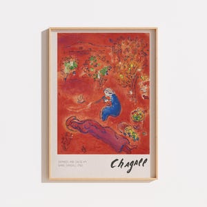 Peut inclure: Une impression d'art encadrée présentant une peinture de Marc Chagall sur fond rouge vif. La scène montre un personnage en bleu jouant de la flûte, un personnage allongé en violet, des arbres et des paniers de fruits. Le texte en bas indique Daphnis and Chloe #4 Marc Chagall 1961.