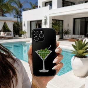 May include: A black smartphone case featuring a decorative graphic of a martini glass filled with green olives. The design is composed of small, sparkling crystals that form the outline of the glass and the round shapes of the olives.