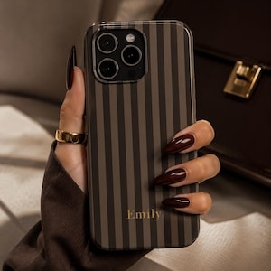 Könnte beinhalten: Eine Smartphone-Hülle mit vertikalem Streifenmuster in abwechselnden Nuancen von Dunkelbraun und Hellbraun. Der Name Emily ist in einer goldenen Serifenschrift unten mittig auf die Hülle gedruckt. Die Hülle hat eine Aussparung für ein Dreifach-Kamerasystem.