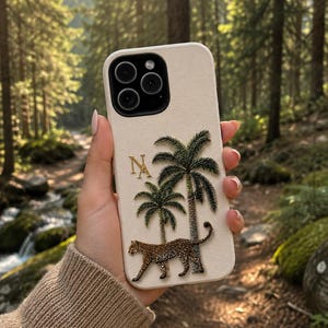 以下が含まれることがあります： クリーム色のスマートフォンケースで、2本の緑色のヤシの木と茶色のヒョウの刺繍が施されています。ケースの左側には金色の文字「NA」があり、上部には黒いカメラ用の切り抜きがあります。