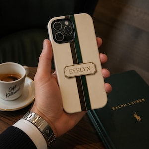 Könnte beinhalten: Eine cremefarbene Smartphone-Hülle mit einem vertikalen dunkelgrünen und braunen Streifen in der Mitte. Eine rechteckige goldfarbene Plakette ist mittig auf dem Streifen angebracht und trägt den Namen EVELYN in serifenbetonten Großbuchstaben.