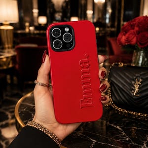 May include: A bright red smartphone case featuring the name Emma embossed in a vertical orientation on the back. The case has a black camera module cutout with three circular lenses and a flash, designed for a modern mobile device.