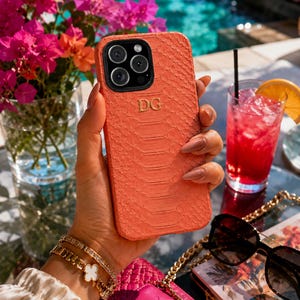 May include: A coral-colored smartphone case with a textured snakeskin pattern and the gold-embossed initials DG centered on the back. The case features a black camera module cutout with three circular lenses and a flash, designed for a modern smartphone.