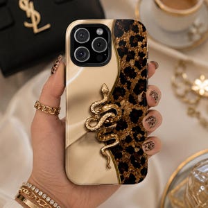 Puede incluir: Una funda para smartphone con un diseño dividido, con una sección beige lisa y sólida a la izquierda y un patrón de leopardo texturizado en marrón y negro a la derecha. Una figura de serpiente de metal dorado está fijada en el centro.