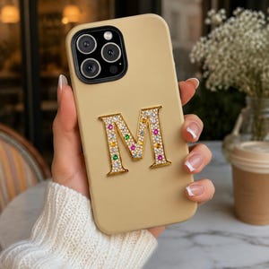 May include: A beige smartphone case featuring a large, gold-toned letter M centered on the back. The letter is encrusted with small, clear white crystals and accented with scattered, colorful gemstones in shades of pink, orange, green, and blue.