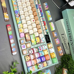 May include: A custom mechanical keyboard with a light green base and colorful keycaps. The keycaps feature various cartoon characters and designs. Additional keycaps are scattered around the keyboard. Two books and some greenery are in the background.