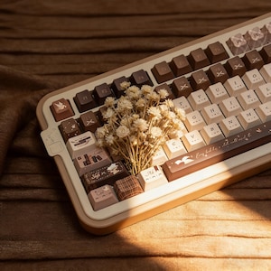 以下が含まれることがあります： ベージュと茶色のカスタムメカニカルキーボード。キーキャップは茶色とベージュの混合色で、一部のキーには装飾的なアイコンが表示されています。小さなドライフラワーの花束がキーボードの上に置かれています。キーボードには「Every day is a new story to tell.」と書かれています。