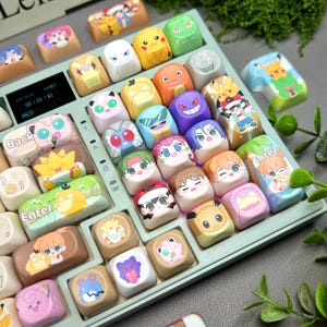 May include: A light green keyboard with custom keycaps featuring various cartoon character designs. The keycaps are colorful and detailed, with characters in shades of yellow, pink, blue, and green. The keyboard has a small digital display.