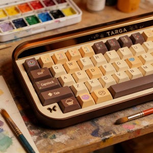 Puede incluir: Teclado mecánico con marco marrón y dorado y teclas blanquecinas. El teclado tiene teclas personalizadas con varios iconos y la palabra "TARGA" impresa en la parte superior. Pinceles y una paleta de acuarela se encuentran en el fondo.