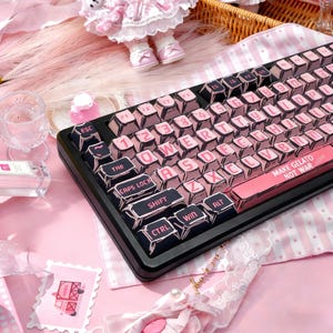 Puede incluir: Un teclado mecánico negro y rosa con teclas rosas y letras negras. Las teclas tienen un contorno de estilo cómic. El teclado tiene el texto "MAKE GELATO NOT WAR" en la barra espaciadora.