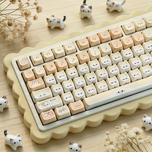 May include: A cream-coloured mechanical keyboard with cat-themed keycaps. The keycaps feature cat faces and other cat-related designs. The keyboard has a scalloped edge and is surrounded by small cat figurines.