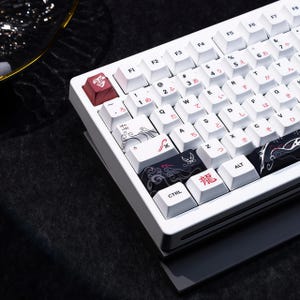 May include: White mechanical keyboard with Japanese characters and decorative keycaps. The keycaps feature red and black designs with Japanese calligraphy and symbols. The keyboard has a white frame and a black base.