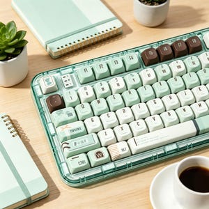 May include: A mint green and white mechanical keyboard with a transparent frame. The keyboard has white and mint green keycaps, with some brown keycaps. A small notebook, succulent plant, and a cup of coffee are also in the image.