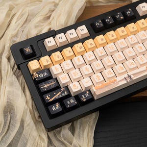 May include: A mechanical keyboard with a black frame and a mix of black and cream-colored keycaps. The cream keys feature gold Chinese characters, while the black keys display gold illustrations of swords, flutes, and additional Chinese text.