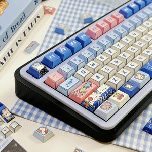May include: A custom mechanical keyboard with pastel-colored keycaps. The keycaps feature a mix of blue, pink, and white colors, with various cute illustrations. The keyboard has a black frame and a light purple base.