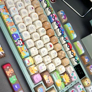 May include: A custom mechanical keyboard with a light green frame and a variety of colorful keycaps. The keycaps feature various designs, including cartoon characters, patterns, and Pokeball designs. The Enter keycap has a green background with cartoon characters and the word "Enter".