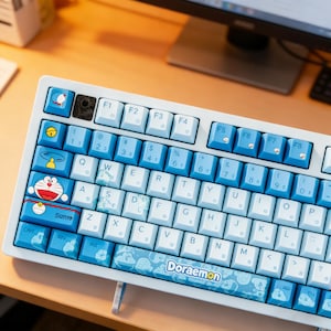 Peut inclure: Un clavier mécanique blanc et bleu avec des touches personnalisées. Les touches présentent le personnage de dessin animé Doraemon et d'autres motifs bleus et blancs. Le clavier possède diverses touches de fonction et un pavé numérique.