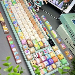 May include: A custom mechanical keyboard with a light green base and a variety of colorful keycaps. The keycaps feature cartoon characters and designs. Additional keycaps are scattered around the keyboard. The keyboard also has a small display screen.