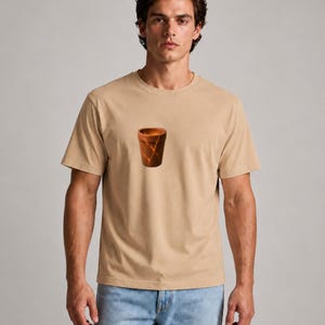 May include: Tan short-sleeve t-shirt featuring a graphic of a brown vase with gold accents. The shirt is made of a soft material and is suitable for casual wear. The model is wearing blue jeans.