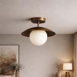 Può includere: Un apparecchio di illuminazione a soffitto con finitura in ottone. L'apparecchio ha una base circolare, uno stelo e un ampio paralume rivolto verso il basso. Un globo di vetro bianco e sferico è sospeso sotto il paralume. La luce è montata su un soffitto di colore neutro.