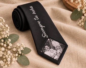May include: A black necktie featuring the white embroidered text "Siempre tu niña" and a rectangular black and white photograph of a person holding a baby, positioned near the pointed end of the tie.