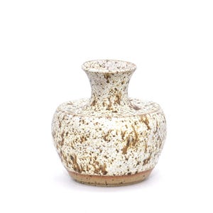 Puede incluir: Un pequeño jarrón de cerámica moteada con cuerpo redondeado y cuello estrecho. El jarrón es principalmente blanco con motas marrones y una base marrón rojiza. El diseño es rústico y hecho a mano.