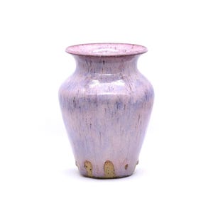 Puede incluir: Un jarrón de cerámica hecho a mano con un suave esmalte lavanda y sutiles acentos azules y marrones. El jarrón tiene un cuerpo redondeado y un borde acampanado, perfecto para exhibir flores o como pieza decorativa. El jarrón mide aproximadamente 18 cm de alto.