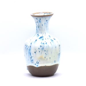 以下が含まれることがあります： 細い首と膨らんだ胴体を持つ陶器製の花瓶。上部は白い釉薬に青い斑点模様が垂れ下がったデザインで、底部は露出した滑らかでマットな茶色の粘土仕上げになっており、円形の縁があります。