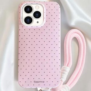 May include: Pale pink phone case with a polka dot design and a matching pink wrist strap. The case has a silver metal ring and the word "Tomorrow" printed on the bottom. The phone case is designed to fit a smartphone.