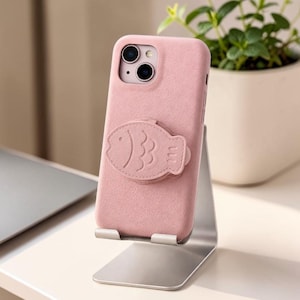 May include: A pink phone case with a fish-shaped grip is displayed on a silver stand. The case has a soft texture and a cutout for the phone's camera. A small plant in a white pot is in the background.