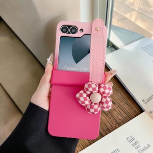 Puede incluir: Una funda de teléfono rosa para un teléfono Galaxy Z Flip. La funda tiene una correa rosa a juego y una flor decorativa con un patrón de cuadros. La pantalla del teléfono muestra las palabras "Galaxy Z Flip".