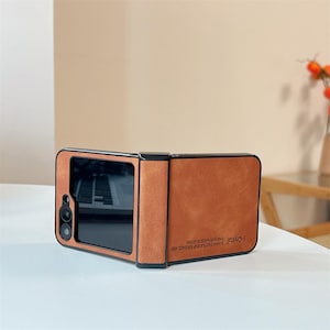 May include: A brown, suede-textured foldable phone case with a black frame and camera cutout. The case features the text i-Crystal A NEW TACTILE EXPERIENCE AND FASHION MADE IN CHINA printed in black on the lower right side of the exterior surface.