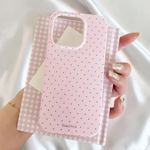 May include: Pink phone case with a polka dot pattern and the word "Tomorrow" printed at the bottom. The case is held over a pink and white checkered notebook.