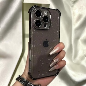 Może przedstawiać: Przezroczyste etui na telefon z brokatem i czarnym zderzakiem. Etui posiada czarne logo Apple i moduł aparatu z czterema obiektywami. Etui na telefon trzymane jest w dłoni z długimi, wypolerowanymi paznokciami.
