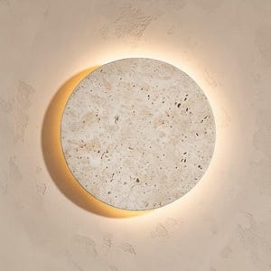 May include: A round, beige wall sconce with a textured surface. The sconce has a warm, glowing light around the edge, creating a halo effect. The sconce is mounted on a light beige wall.