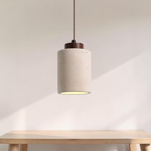 Puede incluir: Una lámpara colgante cilíndrica con una pantalla de hormigón moteado blanquecino y una tapa de madera oscura. La luz cuelga de un cable marrón, iluminando el espacio inferior. La lámpara está suspendida sobre una mesa de madera.