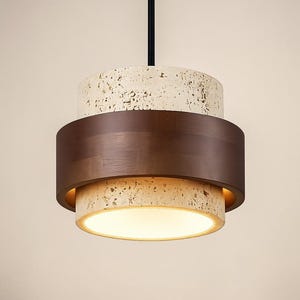Pode incluir: Uma moderna luminária pendente com um design em camadas. Apresenta uma seção superior de material branco-sujo salpicado, uma faixa central de metal marrom escuro e uma seção inferior com uma luz quente. Pendurada por um cabo preto.