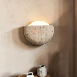 Può includere: Applique da parete beige a forma di semicerchio con superficie strutturata. L'applique emette una luce soffusa e calda. Sotto, sono visibili un piccolo contenitore bianco e un vaso beige con un design circolare.