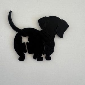 May include: A black dachshund dog silhouette with a star-shaped cutout. The dog is shown in profile, with its tail curved upwards. The cutout is near the dog's rear.