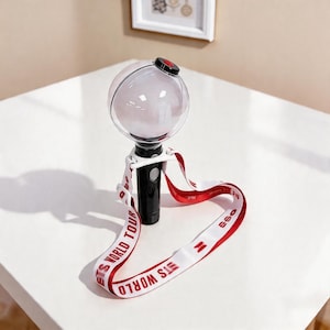 May include: A black and white light stick with a clear orb at the top. The light stick has a red and white strap that says "BTS WORLD TOUR". The light stick is sitting on a white surface.