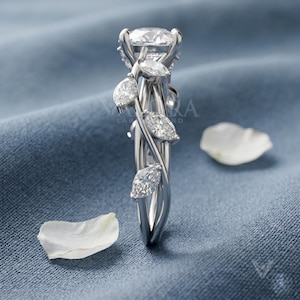 May include: A silver engagement ring with a large, round center stone and smaller, marquise-cut side stones. The ring features a vine-like design with leaf accents. The ring is set against a blue background with white petals.