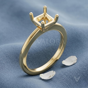 Puede incluir: Un anillo dorado con un engaste cuadrado para una piedra preciosa. El anillo tiene una banda pulida y el texto "VALORA LAB DIAMONDS" es visible. El anillo se muestra sobre una superficie azul.