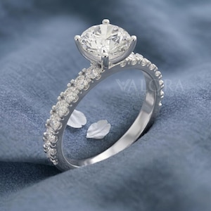 May include: A silver engagement ring featuring a large, round, clear gemstone. The band is set with smaller, clear gemstones. The ring is displayed against a blue backdrop. The text "VALORA LAB GROWN" is visible on the ring.