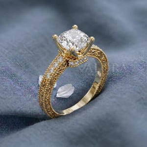 May include: A gold engagement ring with a large, square-cut diamond center stone. The band features intricate filigree and small accent diamonds. The ring is set against a blue background, showcasing its ornate details and sparkling brilliance.