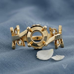 May include: A gold ring featuring a central circular setting with four prongs, flanked by leaf-shaped accents on each side. Each leaf element contains a small, round, clear gemstone. The word VALORA is visible in the background behind the ring.