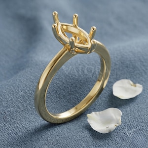 May include: A gold-colored ring setting with a marquise-shaped design, featuring prongs for securing a gemstone. The ring is displayed on a blue surface with two white flower petals. The ring is a jewelry item.
