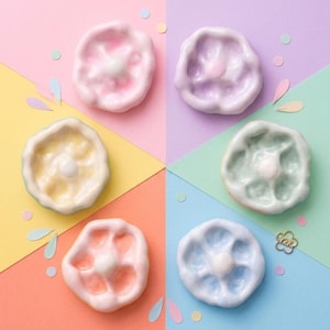 Può includere: Una collezione di sei piatti in ceramica dai colori pastello. Ogni piatto ha un colore unico, tra cui rosa, viola, giallo, verde, pesca e blu. I piatti hanno una forma a fiore con un pomello centrale. Coriandoli decorativi e un ciondolo dorato sparsi.