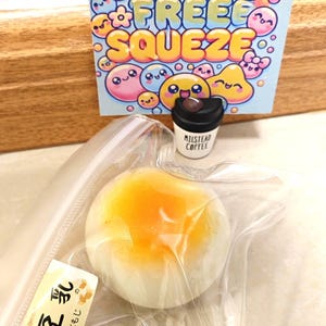 May include: A stress-free squeeze toy set featuring a white and orange squishy bun in a clear bag, a miniature coffee cup, and a colorful sign with the words "STRESS FREE SQUEEZE" and cute cartoon faces.