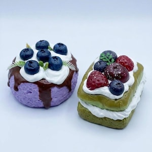 May include: Two decorative food items: a purple cake with blueberries and chocolate drizzle, and a green cake with strawberries and blueberries. Both cakes have white frosting and are on a white surface.
